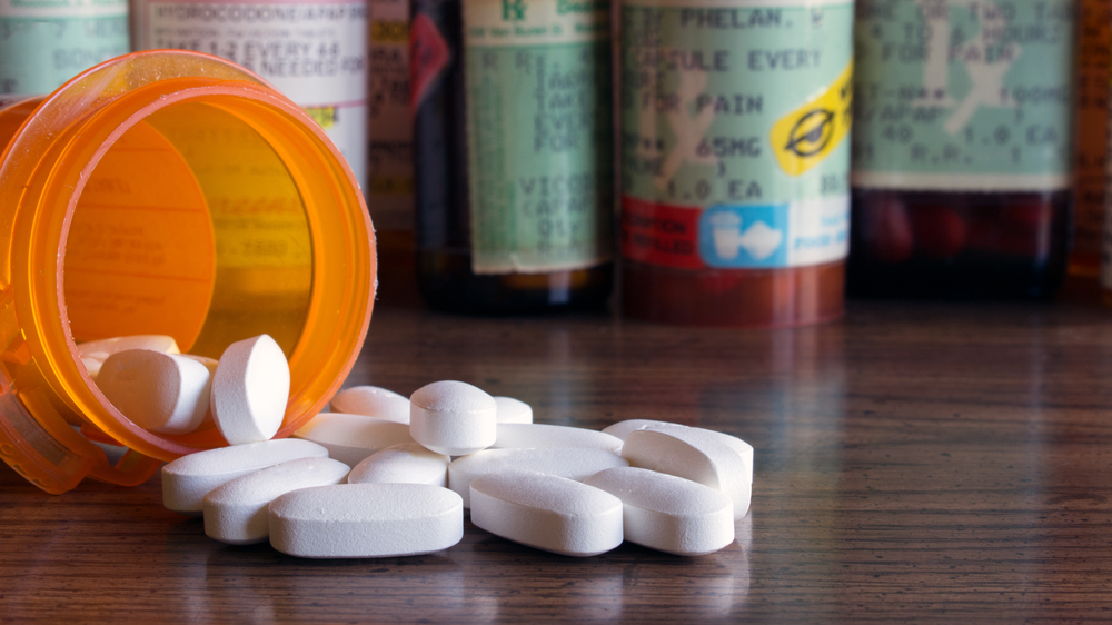 White prescription pills spilled onto a table with many prescription bottles in the background. Concepts of opioid addiction and doctor shopping for prescription pain killers
