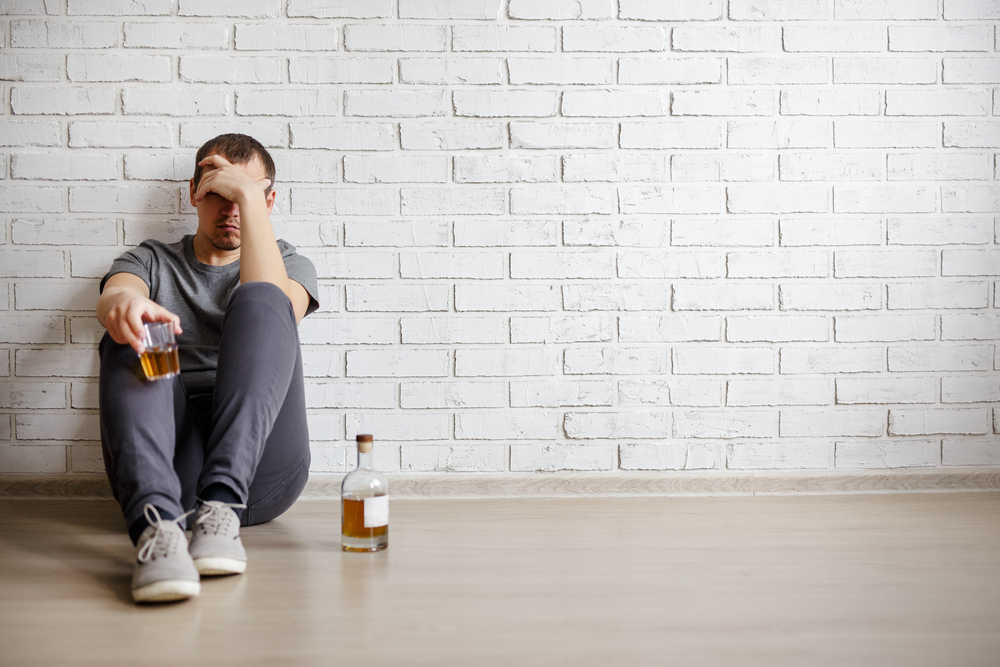 alcoholism concept - sad man with bottle and glass of whiskey sitting on the floor over white brick wall background with copy space