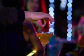 A hand placing GHB in a cocktail at a bar, highlighting addiction risks.