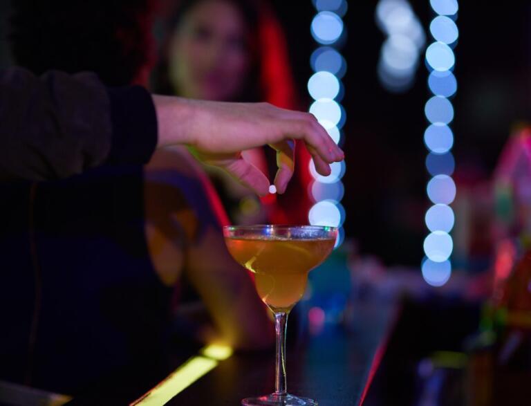 A hand placing GHB in a cocktail at a bar, highlighting addiction risks.