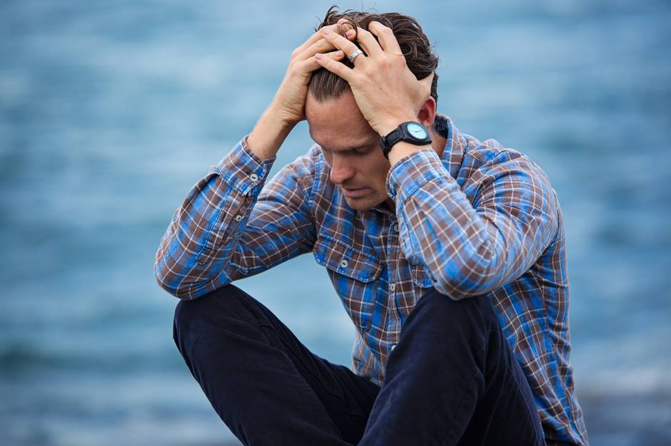 Man holding head in hands by water, reflecting on alcohol recovery