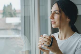 Woman holding coffee mug, looking out window, symbolizing hope in alcohol recovery